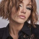 Model wearing a wavy, shoulder-length auburn wig with side parting.